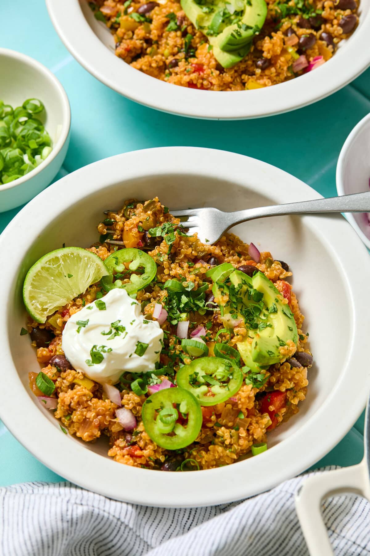 A bowl of quinoa salad topped with sliced avocado, jalapeño, lime wedge, sour cream, chopped red onion, and cilantro, with a fork and a small bowl of green onions nearby.