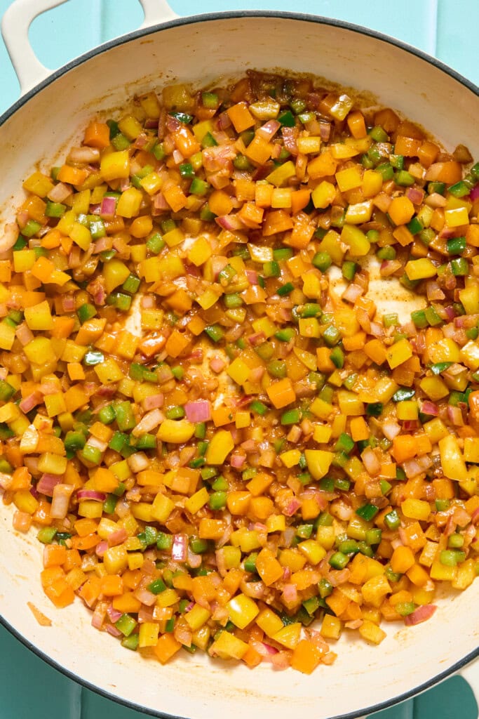 Diced yellow, orange, and green bell peppers with red onions sautéing in a pan, on a light blue surface.