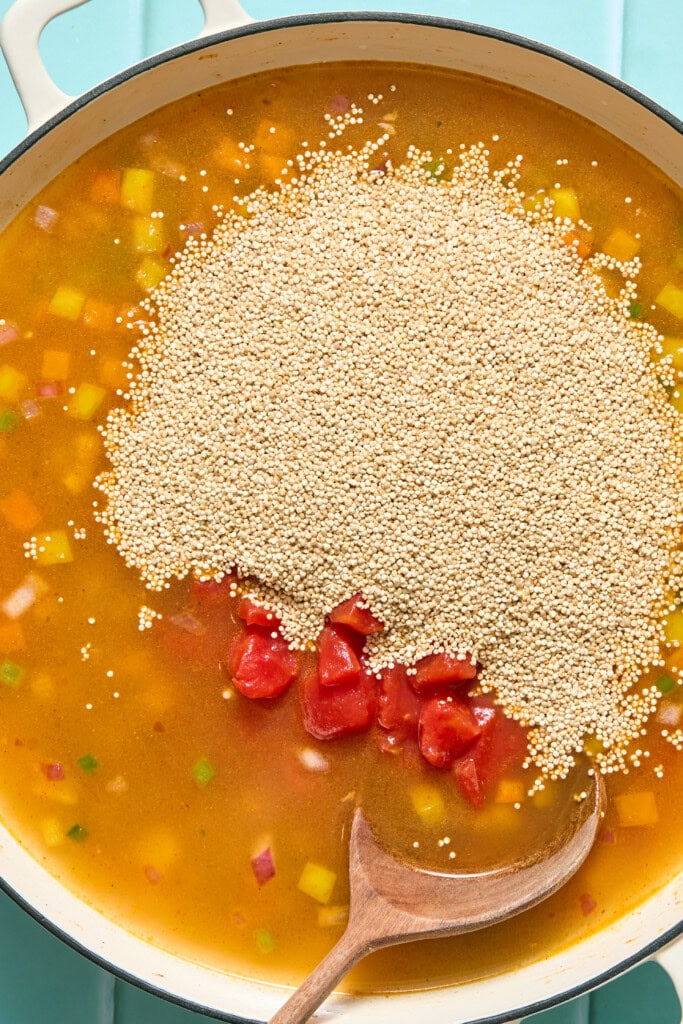 A pot of soup with diced vegetables, broth, chopped tomatoes, and a large pile of uncooked quinoa on top. A wooden spoon rests in the pot.