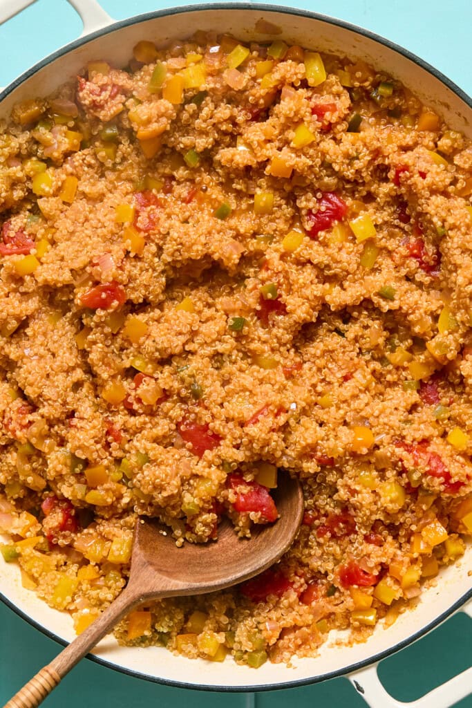 A skillet filled with cooked quinoa mixed with diced yellow peppers, tomatoes, and green herbs, with a wooden spoon resting in the mixture on a light blue surface.