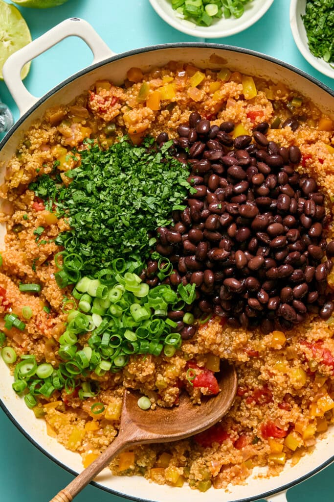 A skillet filled with a quinoa and vegetable mixture, topped with black beans, chopped green onions, and fresh cilantro, with a wooden spoon resting inside. Bowls of green onions and limes are visible in the background.