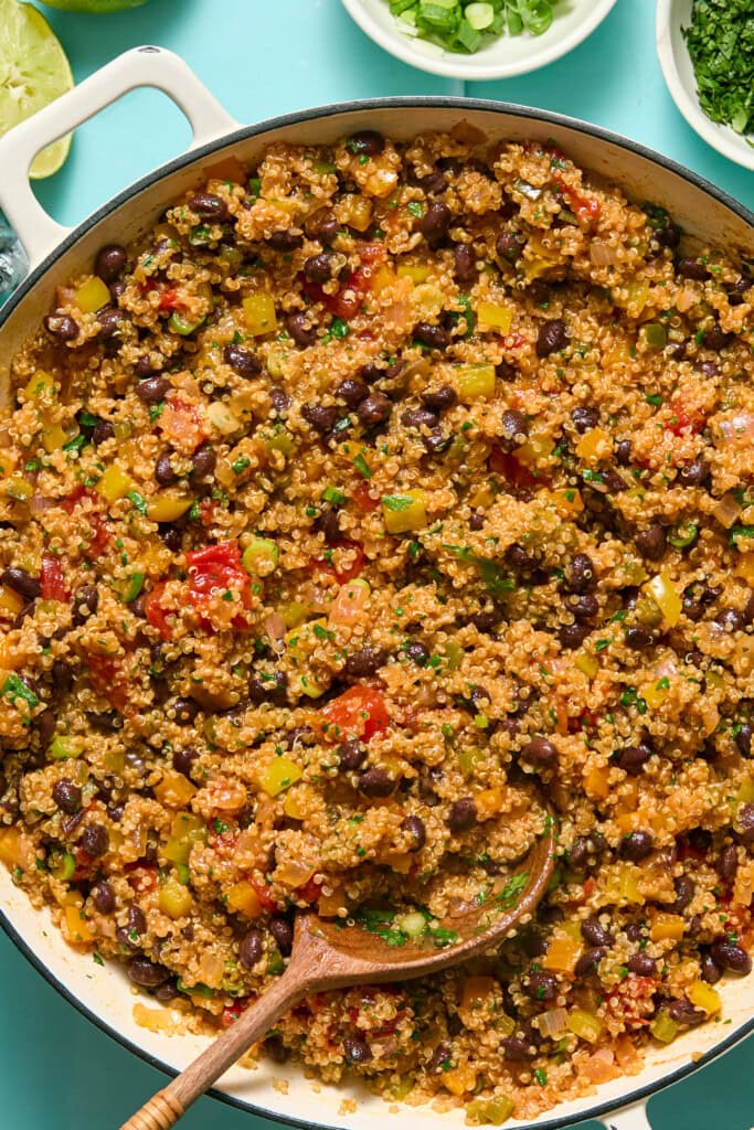 A skillet filled with quinoa, black beans, chopped bell peppers, tomatoes, and spices, with a wooden spoon resting inside. Small bowls of green onions and herbs are visible in the background.