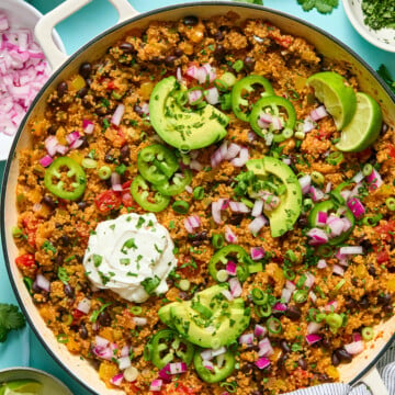A skillet of quinoa and black bean taco filling topped with sliced avocado, jalapeño, lime wedges, diced red onion, green onions, cilantro, and a dollop of sour cream, with garnish bowls nearby.