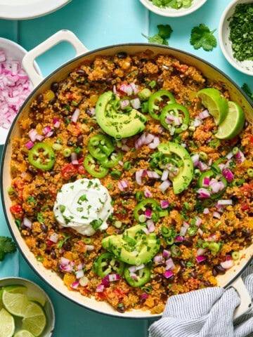 A skillet of quinoa and black bean taco filling topped with sliced avocado, jalapeño, lime wedges, diced red onion, green onions, cilantro, and a dollop of sour cream, with garnish bowls nearby.