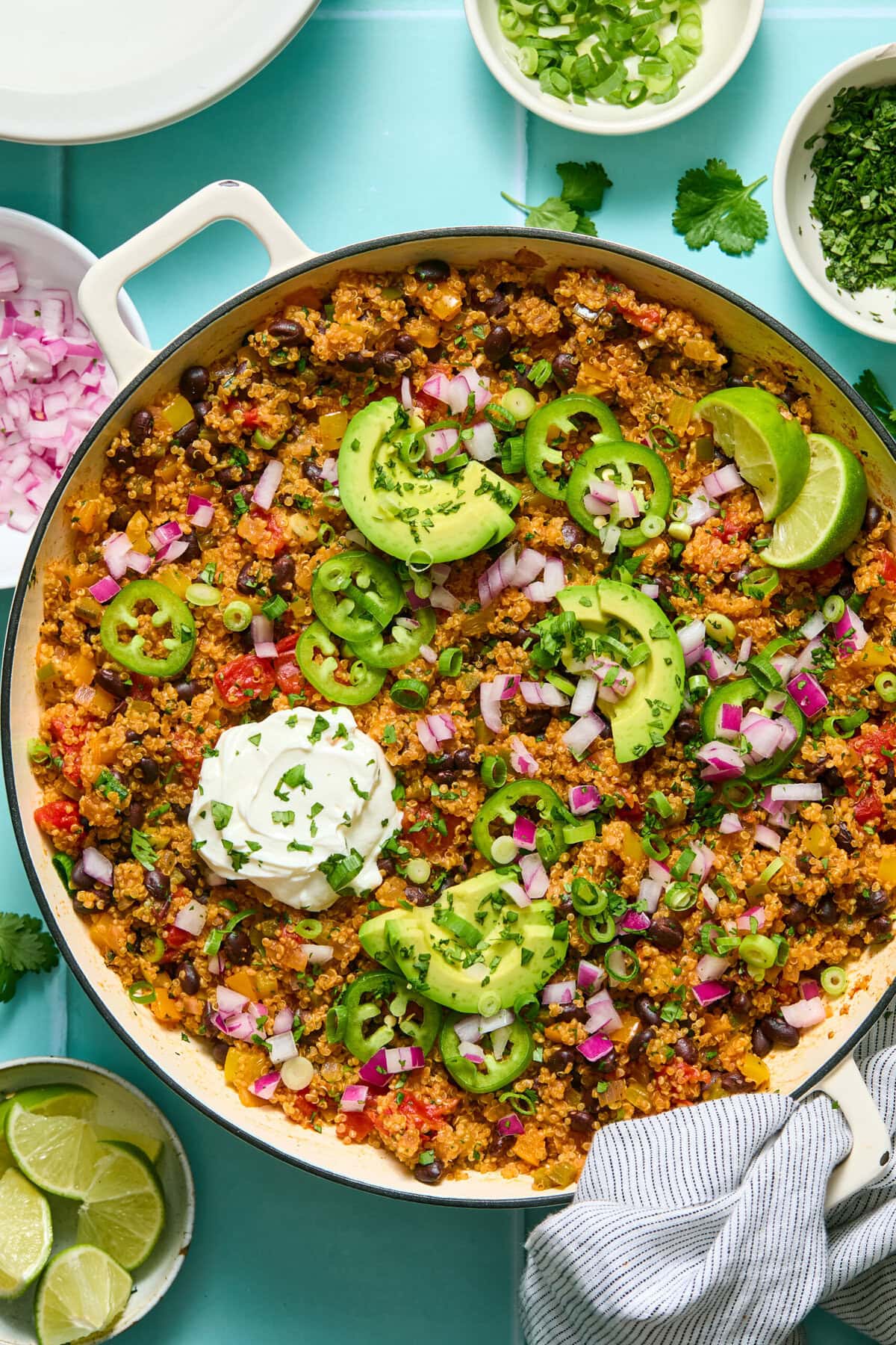 A skillet of quinoa and black bean taco filling topped with sliced avocado, jalape&ntilde;o, lime wedges, diced red onion, green onions, cilantro, and a dollop of sour cream, with garnish bowls nearby.
