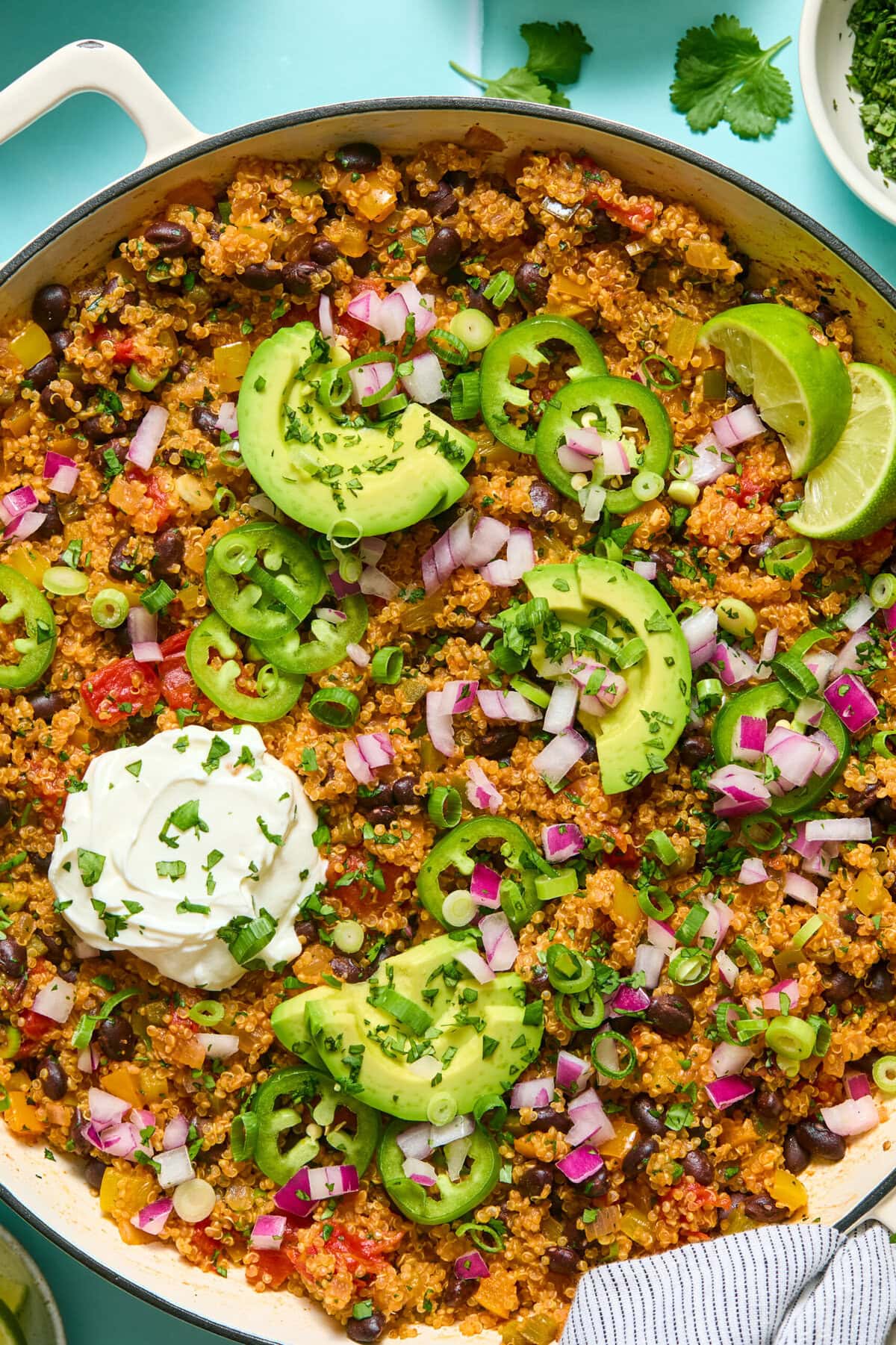 A skillet of quinoa and black bean casserole topped with sliced avocado, jalapeños, chopped red onion, green onion, fresh cilantro, lime wedges, and a dollop of sour cream.