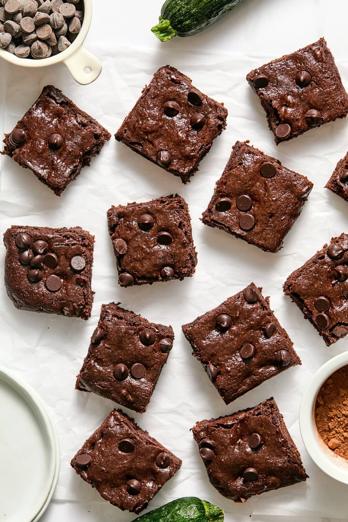 Twelve decadent zucchini brownies with chocolate chips are arranged on white parchment paper. A bowl of chocolate chips, a fresh zucchini, and a small plate are nearby.