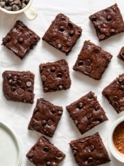 Twelve decadent zucchini brownies are arranged on white parchment paper, with bowls of chocolate chips and cocoa powder nearby.
