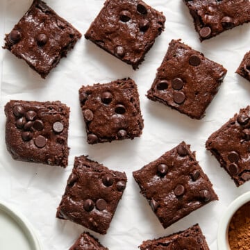 Twelve decadent zucchini brownies are arranged on white parchment paper, with bowls of chocolate chips and cocoa powder nearby.