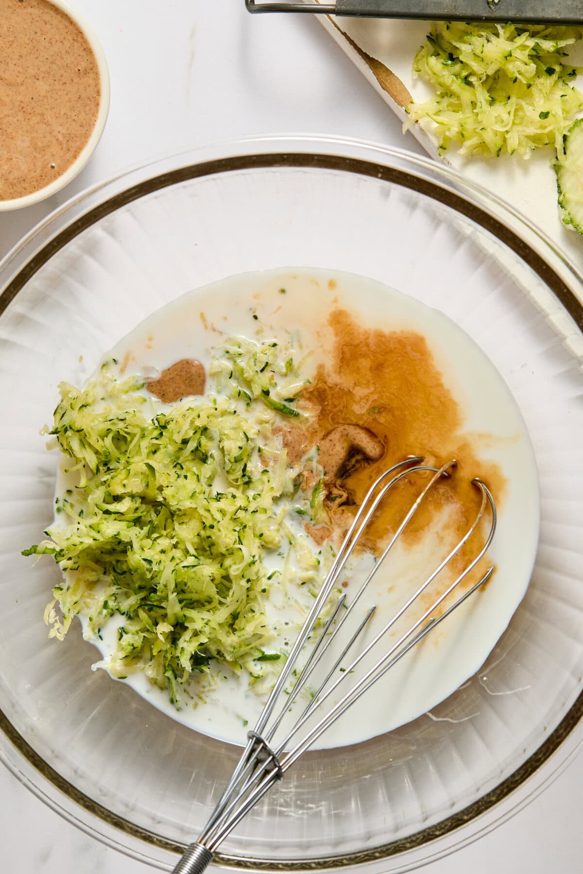 A glass bowl with shredded zucchini, milk, vanilla, and cinnamon being mixed with a whisk-perfect for whipping up moist and flavorful zucchini brownies; grated zucchini and a small bowl of cinnamon rest on the side.