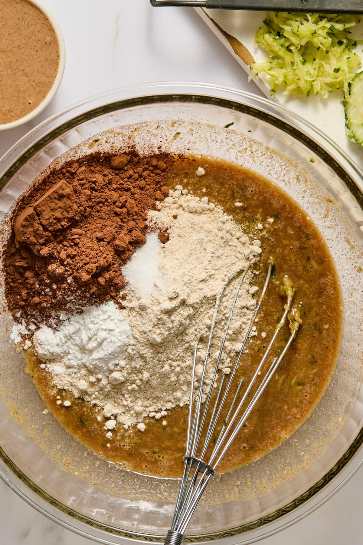 A glass bowl with a whisk, filled with wet ingredients, cocoa powder, flour, and baking powder for zucchini brownies. Shredded zucchini and a small bowl of brown liquid are nearby on a marble surface.