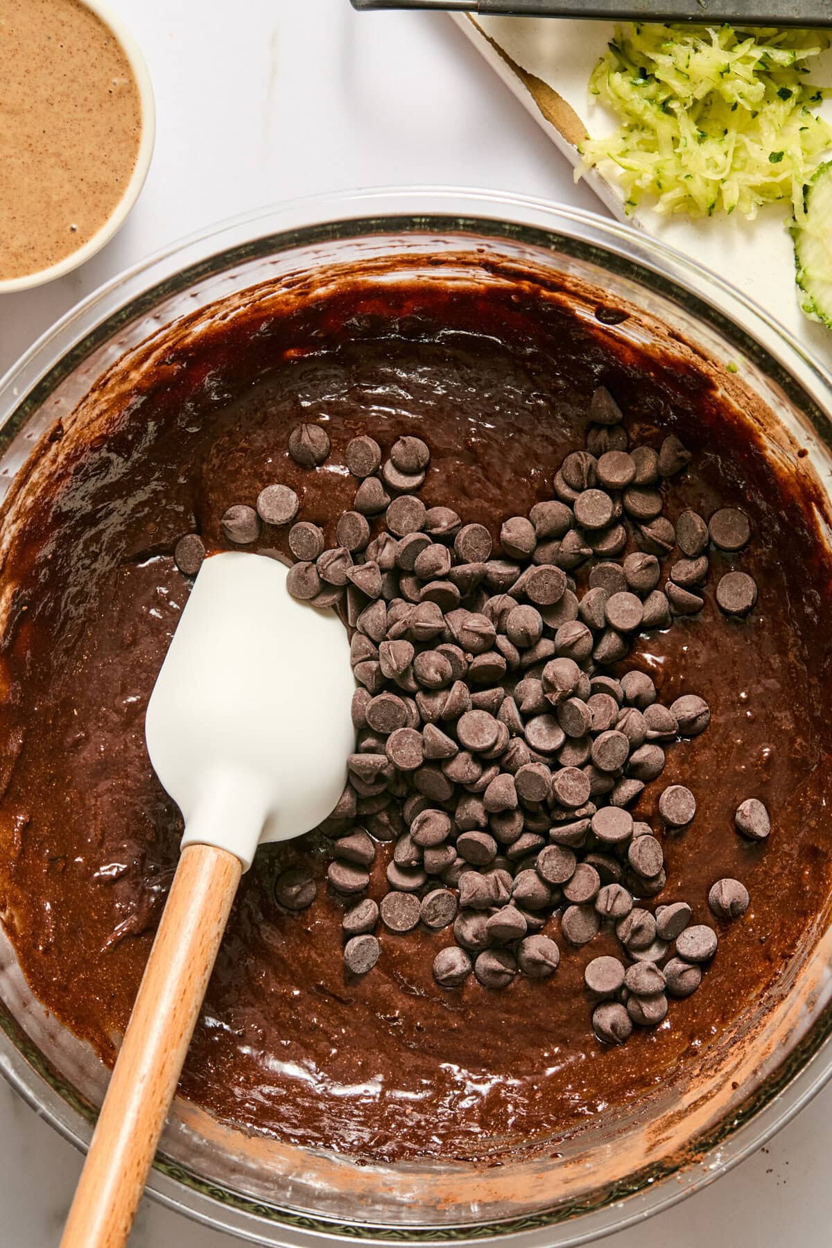 A bowl of chocolate batter with chocolate chips and a white spatula, perfect for making zucchini brownies. There is shredded zucchini and a small bowl of brown liquid nearby on the table.
