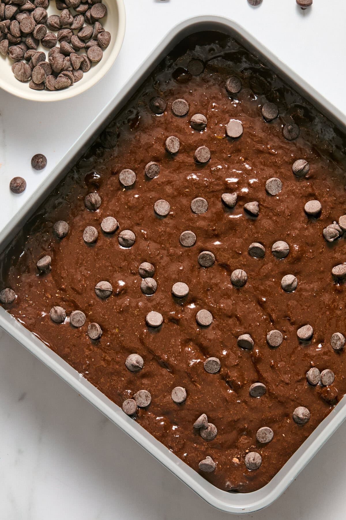 A square baking pan filled with unbaked zucchini brownie batter topped with chocolate chips sits on a white countertop. A small bowl of extra chocolate chips is nearby.