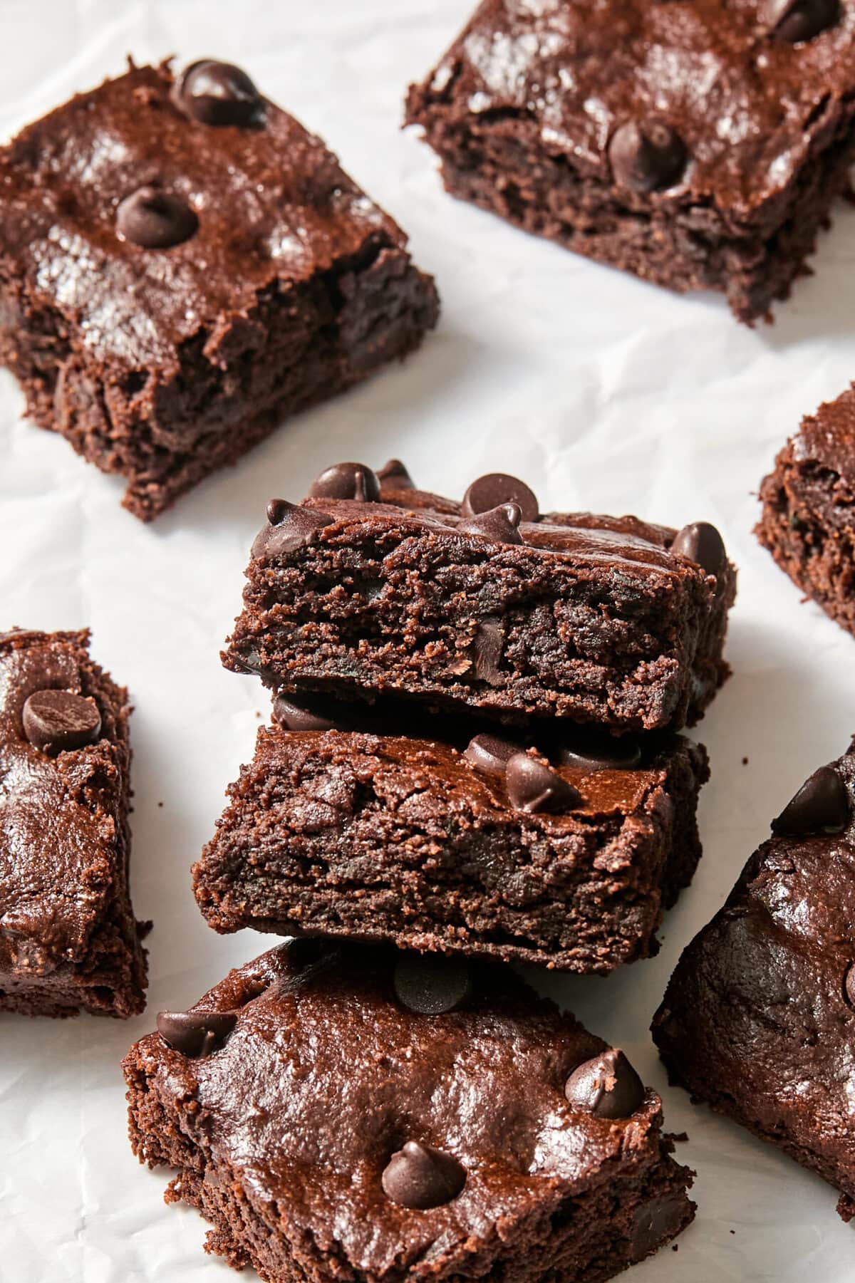 Fudgy chocolate zucchini brownies with chocolate chips are stacked and scattered on white parchment paper. The brownies have a rich, moist texture and glossy tops.