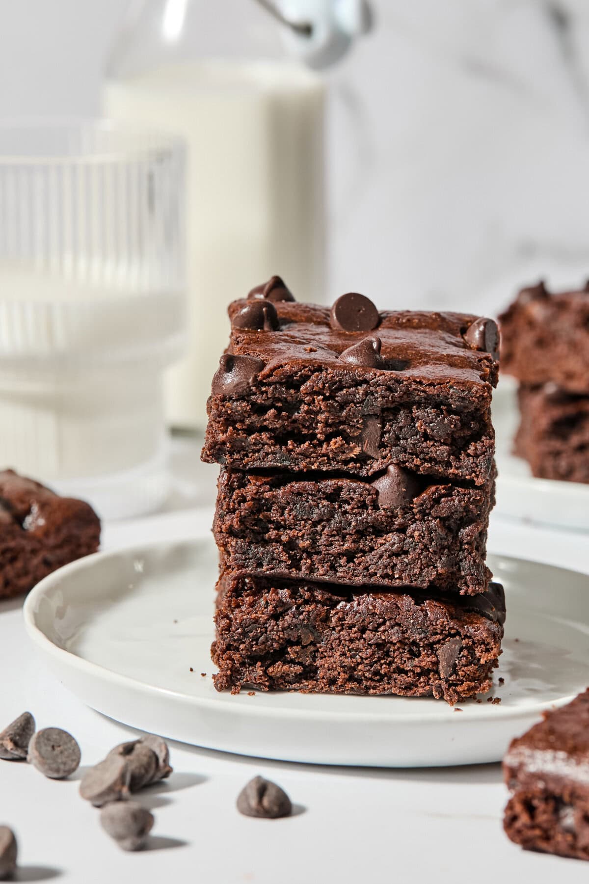 A stack of three rich zucchini brownies with chocolate chips sits on a white plate, with a glass and bottle of milk in the background and scattered chocolate chips nearby.