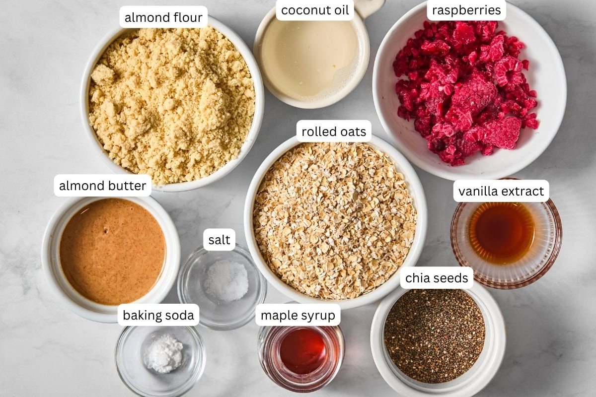 Bowls of almond flour, coconut oil, raspberries, rolled oats, almond butter, salt, baking soda, maple syrup, vanilla extract, and chia seeds on a white surface-perfect for creating delicious vegan almond butter & jelly bars-with each ingredient clearly labeled.