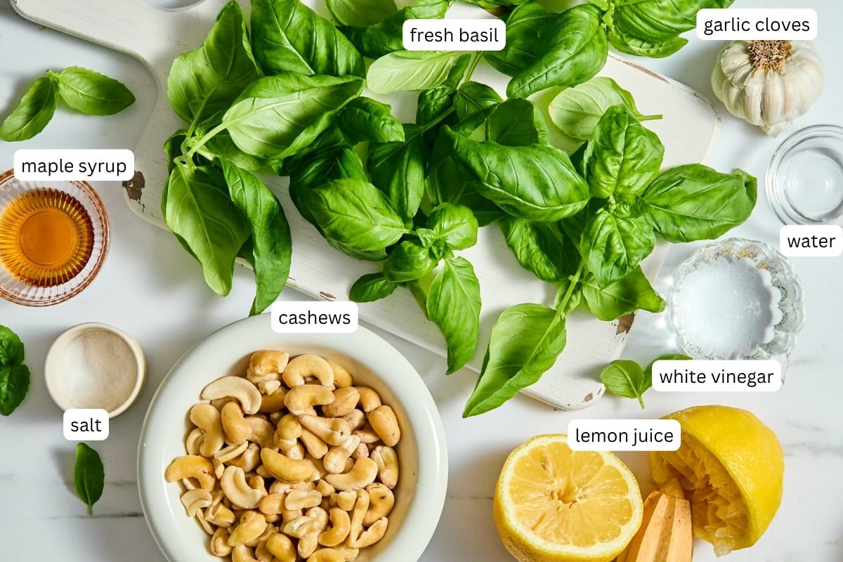 A spread of ingredients labeled: fresh basil, garlic cloves, water, white vinegar, lemon juice, cashews, salt, and maple syrup-perfect for making a creamy dairy-free basil sauce-arranged on a white surface with a bowl of cashews and halved lemon.