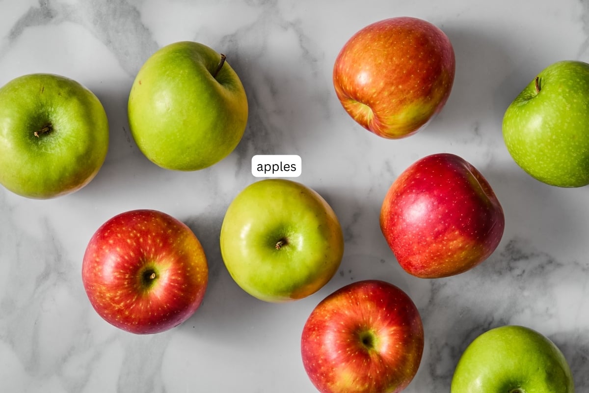 A mix of green and red apples are arranged on a white marble surface, hinting at the perfect ingredients for crispy apple chips made with no dehydrator. A small label in the center reads apples.