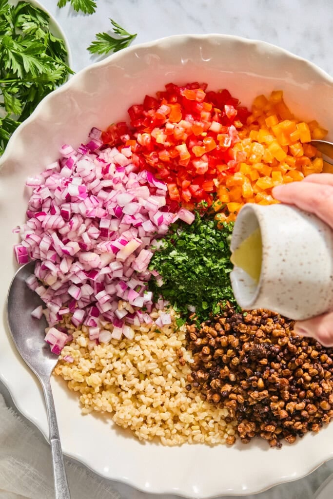 A hand pours dressing onto a bowl filled with diced red onion, tomatoes, yellow bell pepper, parsley, bulgur, and roasted chickpeas. A spoon rests on the bowls edge. Fresh herbs are visible nearby.