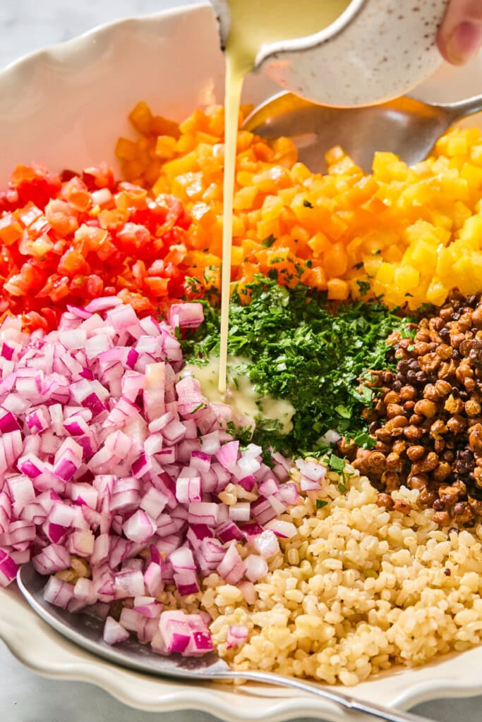 A bowl with chopped red onion, red and yellow bell peppers, parsley, roasted chickpeas, and brown rice, with dressing being poured on top.