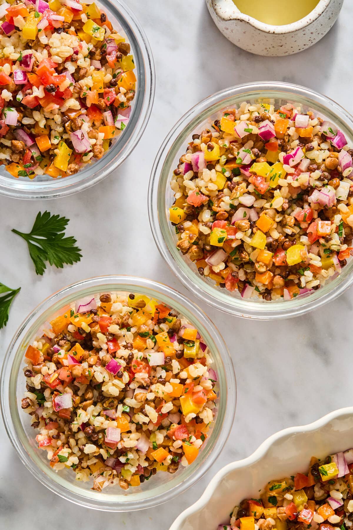 Four glass bowls filled with colorful chopped salad, including brown rice, lentils, red onions, bell peppers, and herbs, are arranged on a marble surface near a small bowl of dressing and parsley leaves.