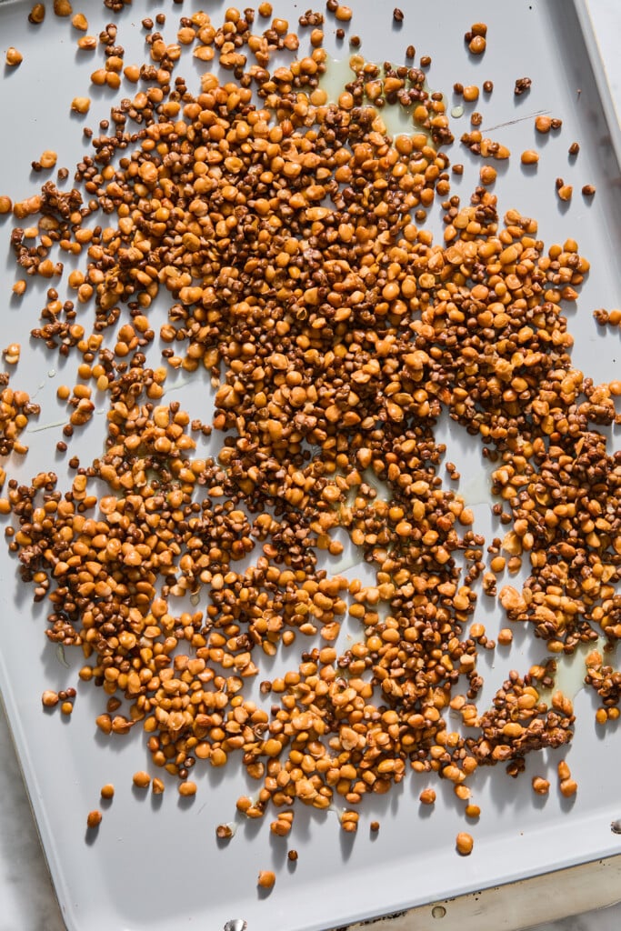 Toasted chickpeas scattered on a light-colored baking tray, some pieces darker and unevenly roasted, with small patches of oil visible.