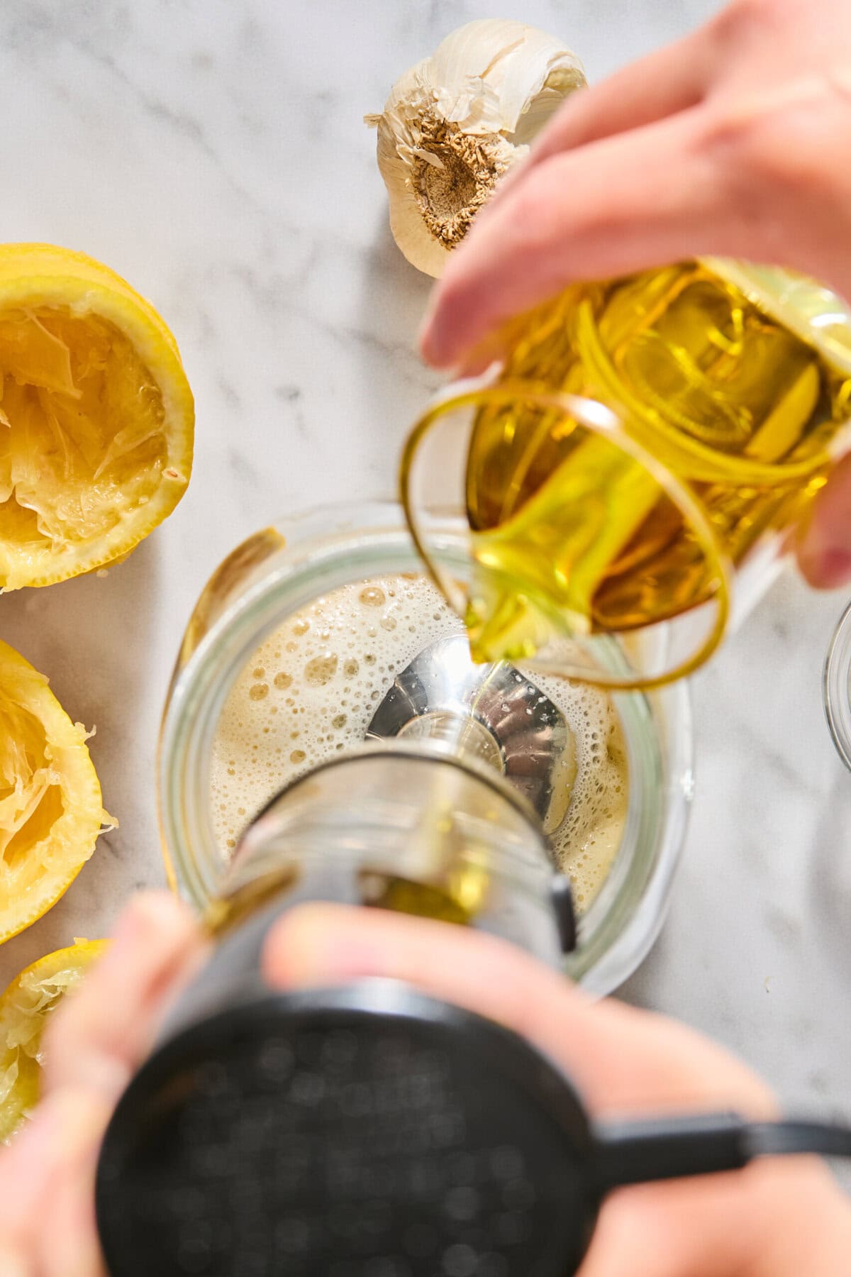 A hand pours olive oil into a blender with lemon halves and a garlic bulb nearby on a marble surface, preparing ingredients for a recipe.