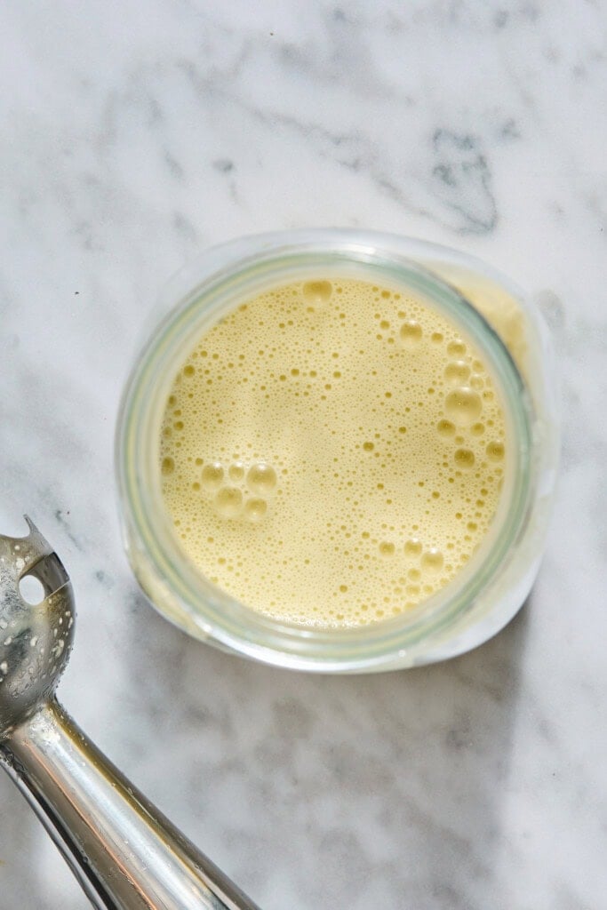 A jar filled with frothy, light yellow liquid sits on a marble surface next to a metal immersion blender with some liquid residue.