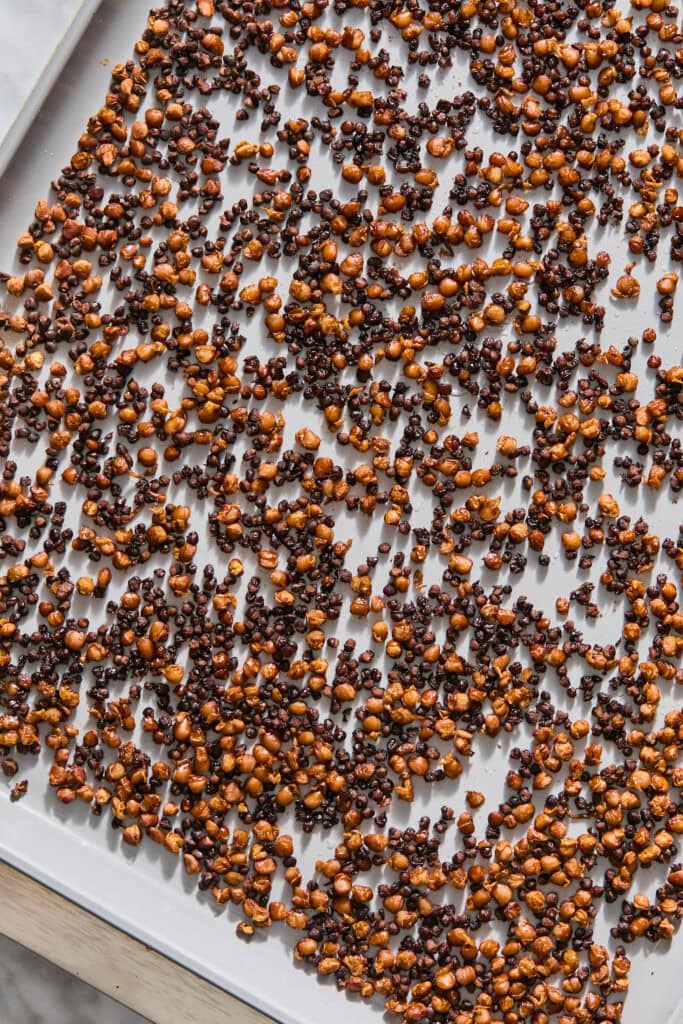 A baking sheet covered with roasted, crispy brown and black quinoa kernels, spread out evenly on a light-colored parchment paper.