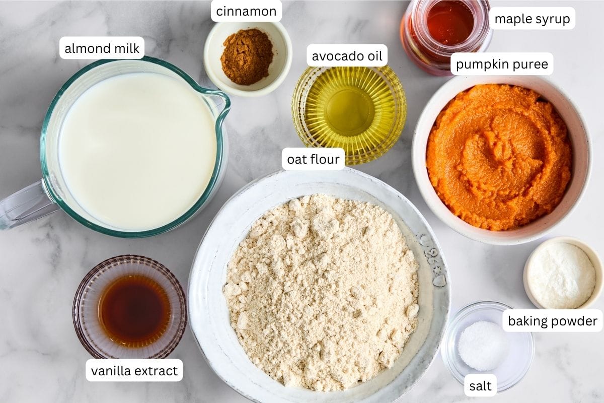 Various labeled ingredients in small bowls on a white surface-almond milk, cinnamon, avocado oil, maple syrup, pumpkin puree, oat flour, vanilla extract, baking powder, and salt-all come together to create delicious Vegan Pumpkin Pancakes. Top-down view.