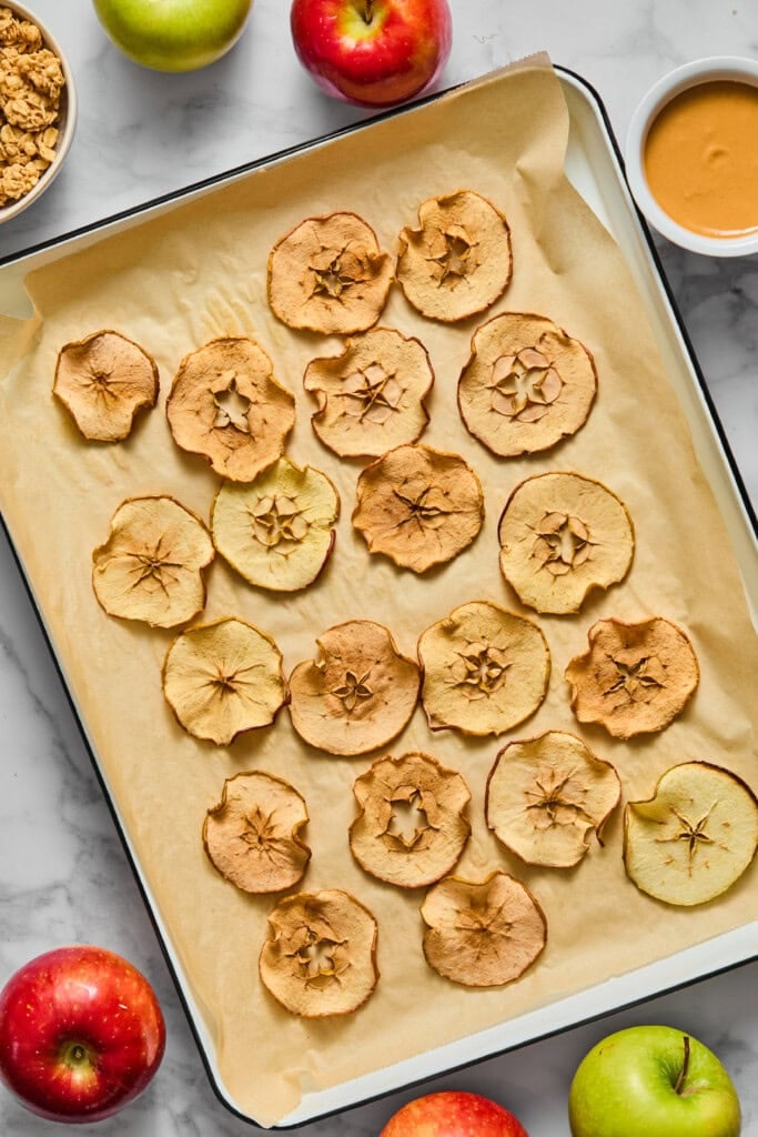 A baking tray with crispy apple chips on parchment paper, surrounded by fresh apples, a bowl of granola, and a bowl of peanut butter on a marble surface.
