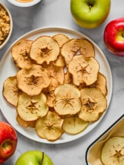 A plate of crispy apple chips on a white surface, surrounded by whole green and red apples, a bowl of granola, a dish of peanut butter, and a tray lined with parchment paper.