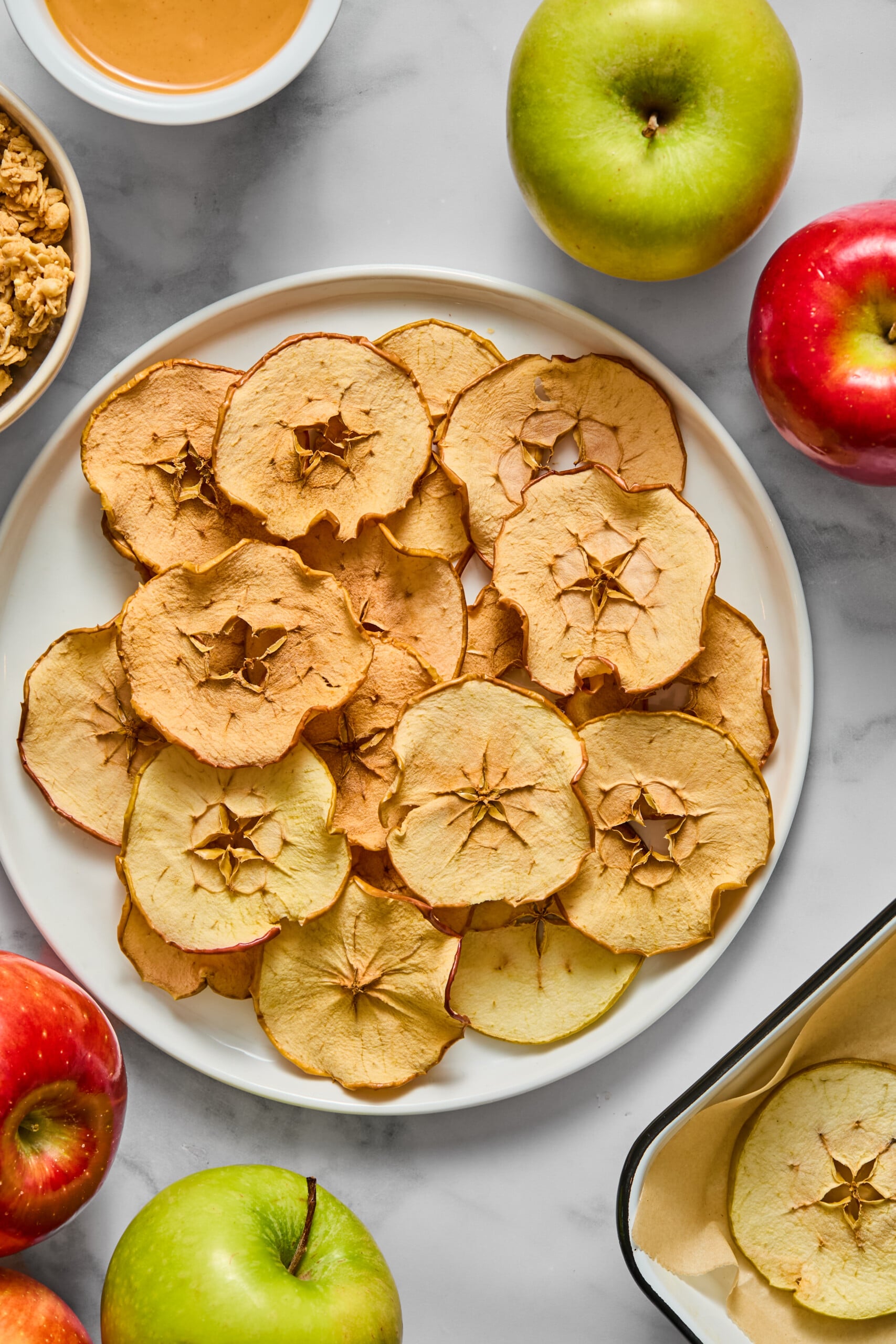 A plate of crispy apple chips on a white surface, surrounded by whole green and red apples, a bowl of granola, a dish of peanut butter, and a tray lined with parchment paper.