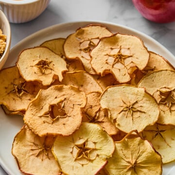 A white plate of crispy apple chips sits on a table, surrounded by whole red and green apples, a bowl of caramel dip, and another bowl of granola.