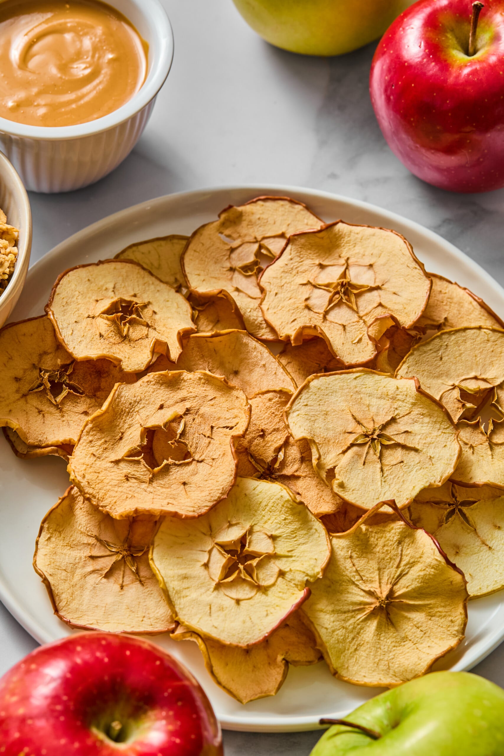 A white plate of crispy apple chips sits on a table, surrounded by whole red and green apples, a bowl of caramel dip, and another bowl of granola.
