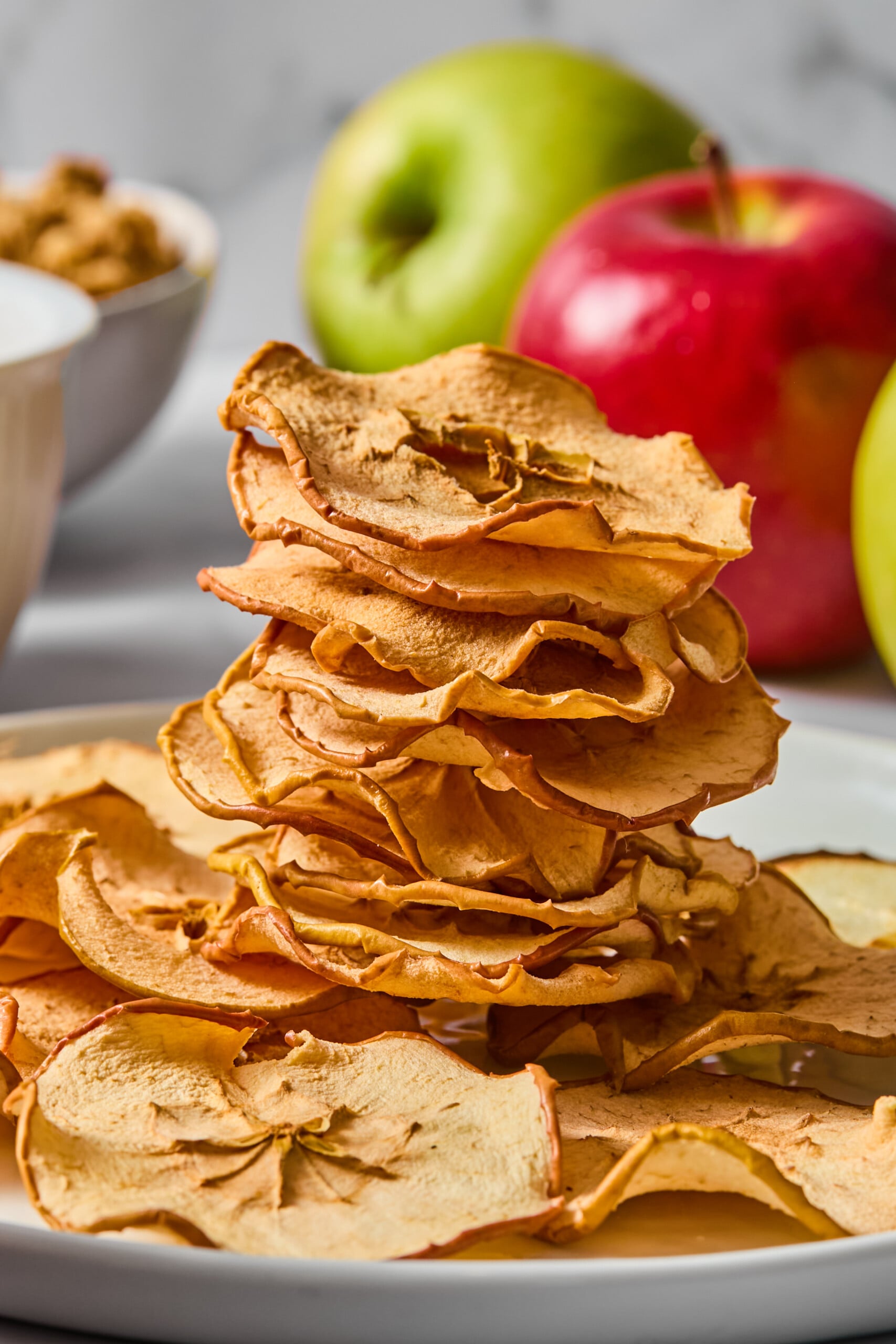 A stack of crispy apple chips rests on a white plate, with fresh green and red apples and a bowl of granola in the blurred background.
