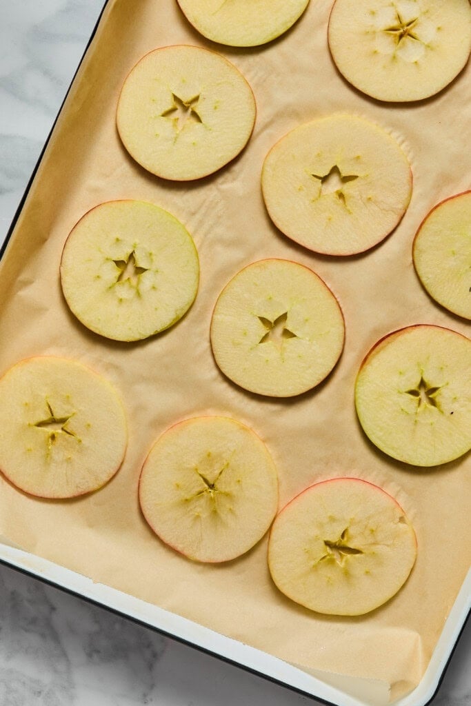 Thin apple slices with visible star-shaped cores are arranged in neat rows on a parchment-lined baking sheet, ready for baking or drying.