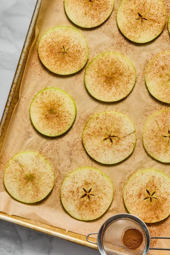 Thin apple slices sprinkled with cinnamon are arranged on a parchment-lined baking sheet. A small mesh sieve with cinnamon sits in the lower right corner on the tray.
