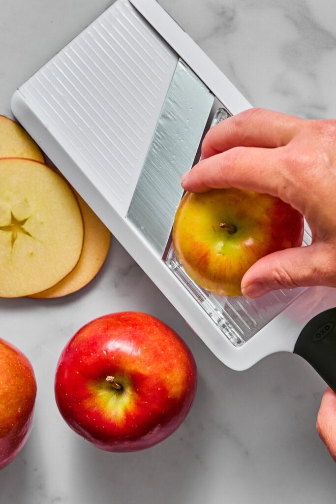 A hand slices an apple with a white mandoline slicer on a marble surface. Two whole apples and several thin apple slices are nearby.