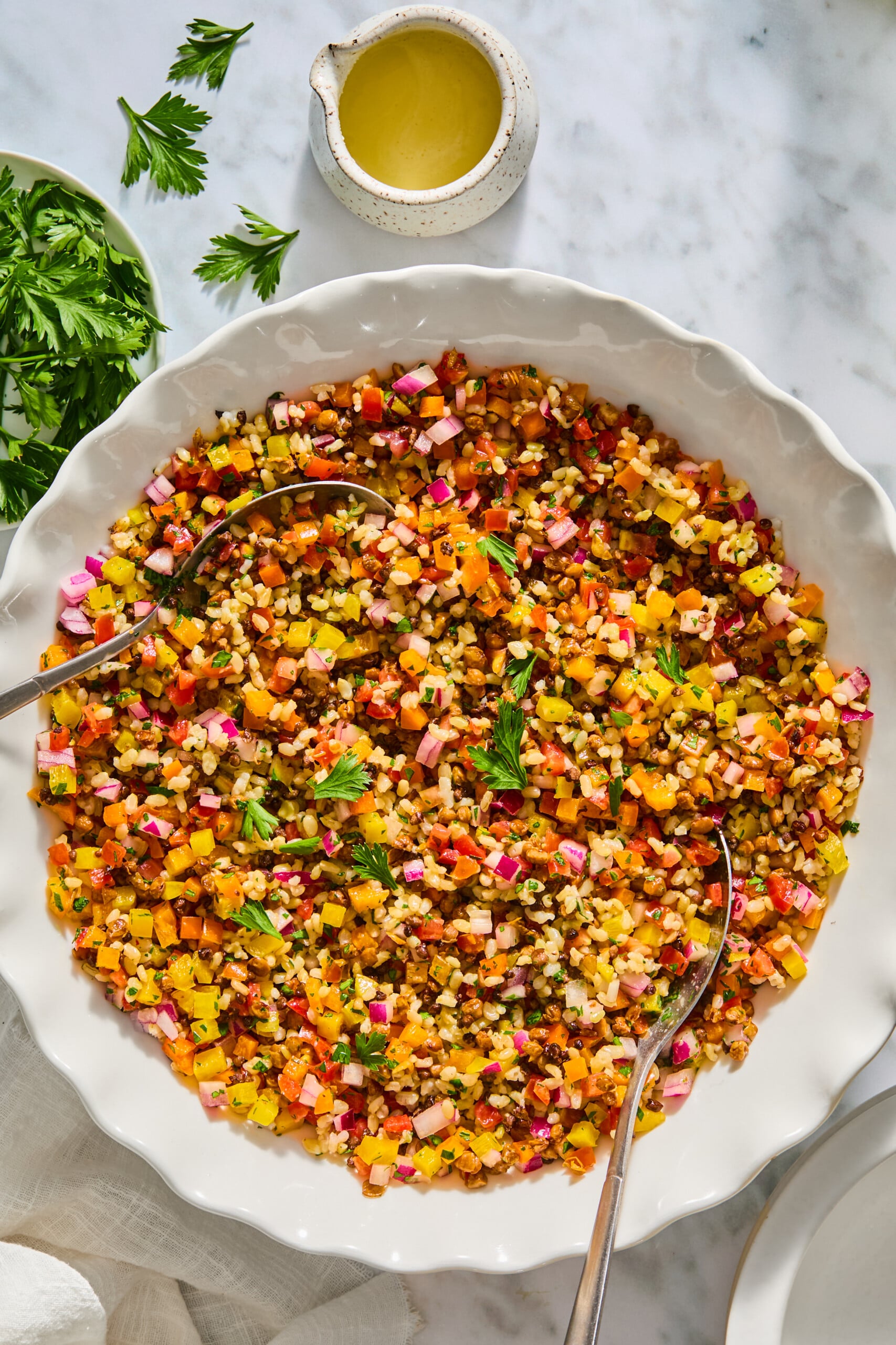 A large white bowl filled with finely chopped colorful vegetables and grains creates a vibrant lentil rice salad, with two serving spoons ready for sharing. Fresh parsley and a small dish of dressing are nearby on the marble surface.