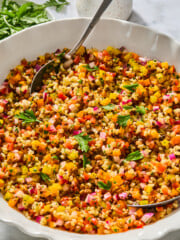A bowl of colorful bulgur and lentil rice salad with finely chopped vegetables, garnished with fresh parsley, and a serving spoon resting inside. A small jug and fresh herbs are in the background.
