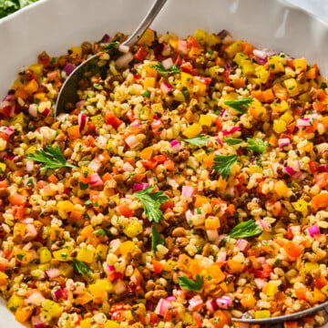 A bowl of colorful bulgur and lentil rice salad with finely chopped vegetables, garnished with fresh parsley, and a serving spoon resting inside. A small jug and fresh herbs are in the background.