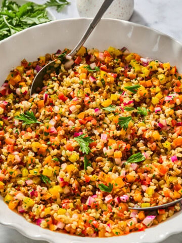 A bowl of colorful bulgur and lentil rice salad with finely chopped vegetables, garnished with fresh parsley, and a serving spoon resting inside. A small jug and fresh herbs are in the background.