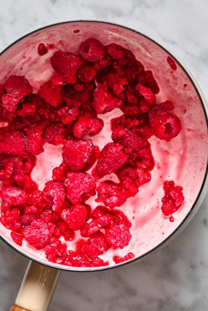 A saucepan with partially melted frozen raspberries mixed with cream or yogurt sits on a marble countertop.