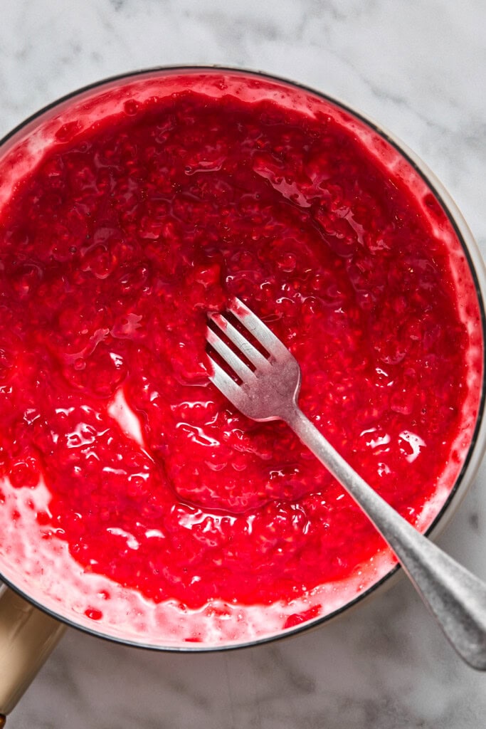 A metal pan filled with bright red raspberry sauce, reminiscent of the sweet filling in almond butter and jelly bars, is being stirred with a silver fork and shown on a white marble surface.