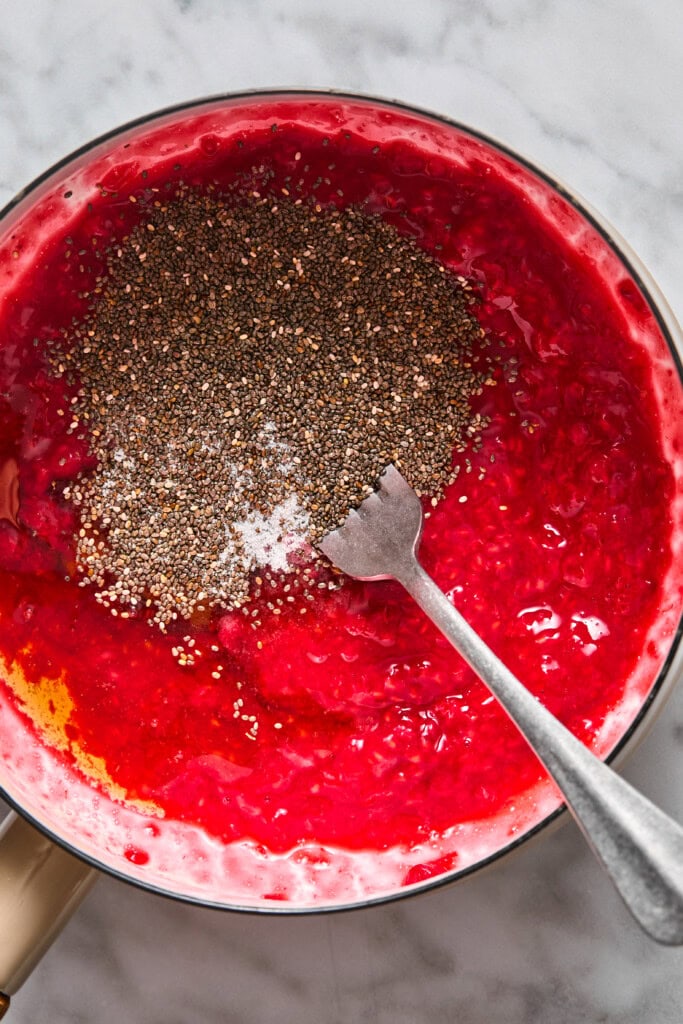 A metal fork rests in a bowl of bright red fruit mixture with chia seeds sprinkled on top, ready to be mixed, reminiscent of the vibrant flavors found in almond butter and jelly bars, all set on a marble tabletop.
