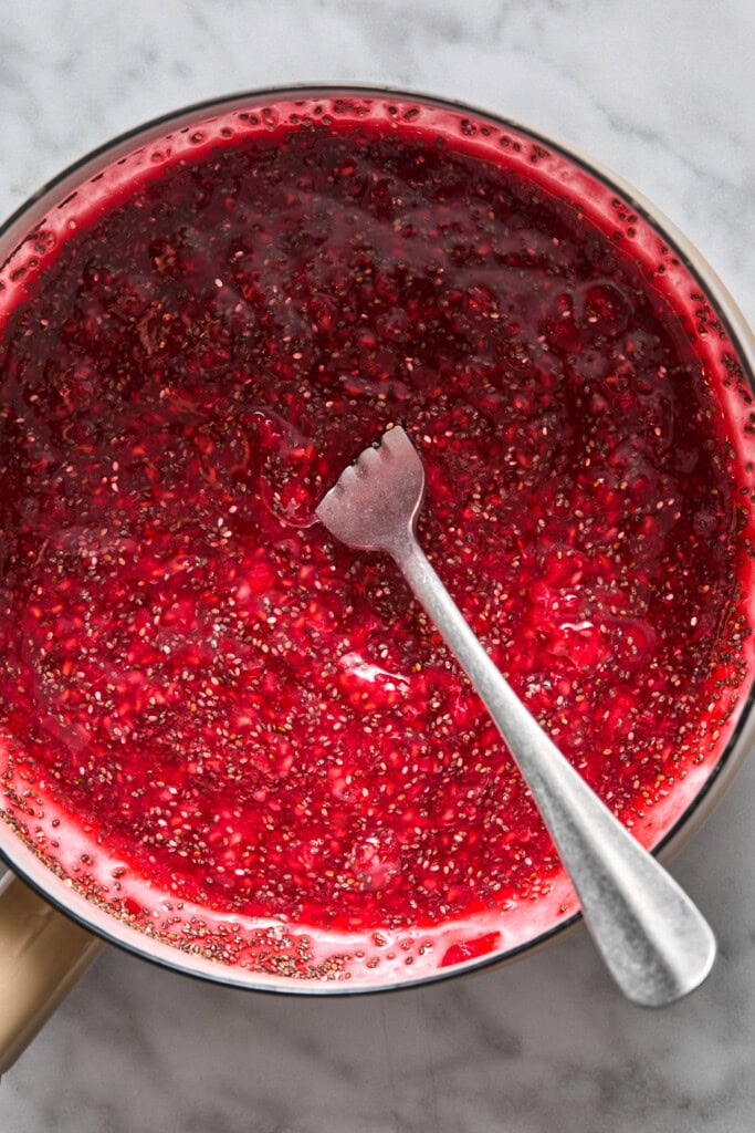 A silver fork stirs a thick, red raspberry and chia seed mixture in a saucepan on a marble countertop—an essential step for preparing delicious almond butter and jelly bars.