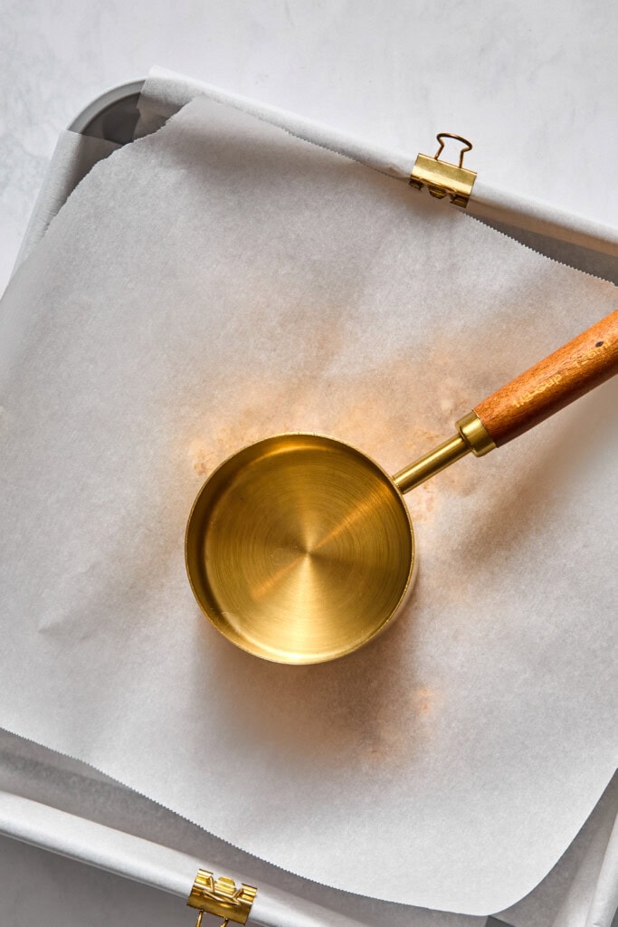 A gold measuring cup with a wooden handle rests on parchment paper in a baking tray, secured with two binder clips at the corners-perfect for preparing vegan almond butter jelly bars.
