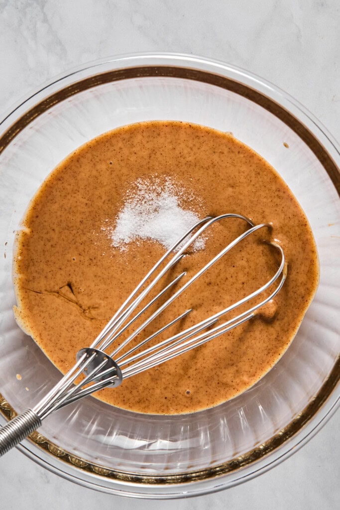 A glass bowl with brown almond butter batter, a pile of white salt on top, and a metal whisk resting inside, on a light surface-perfect for making delicious vegan jelly bars.