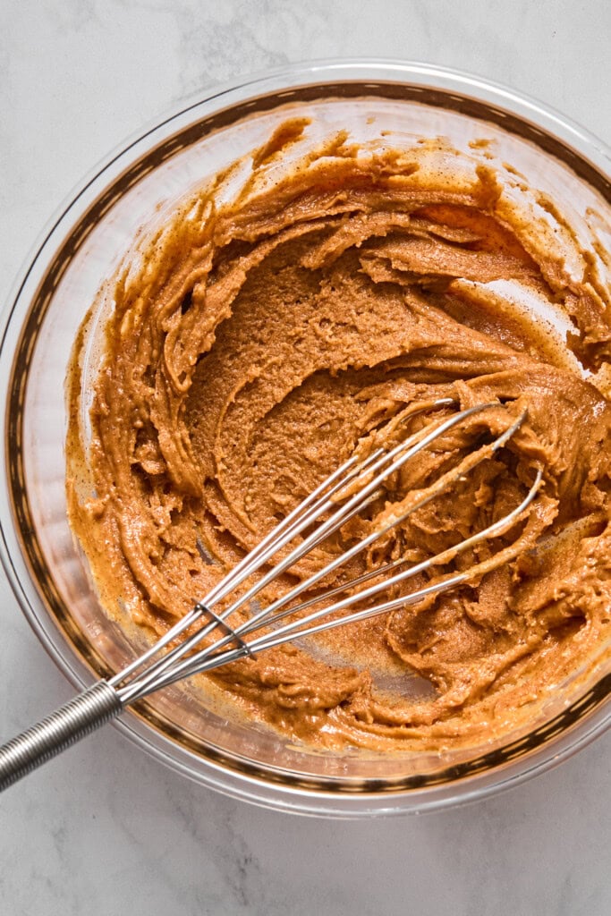 A clear glass bowl sitting on a white marble surface contains a thick, creamy mixture of light brown vegan batter being stirred by a metal wire whisk, perfect for making almond butter jelly bars.